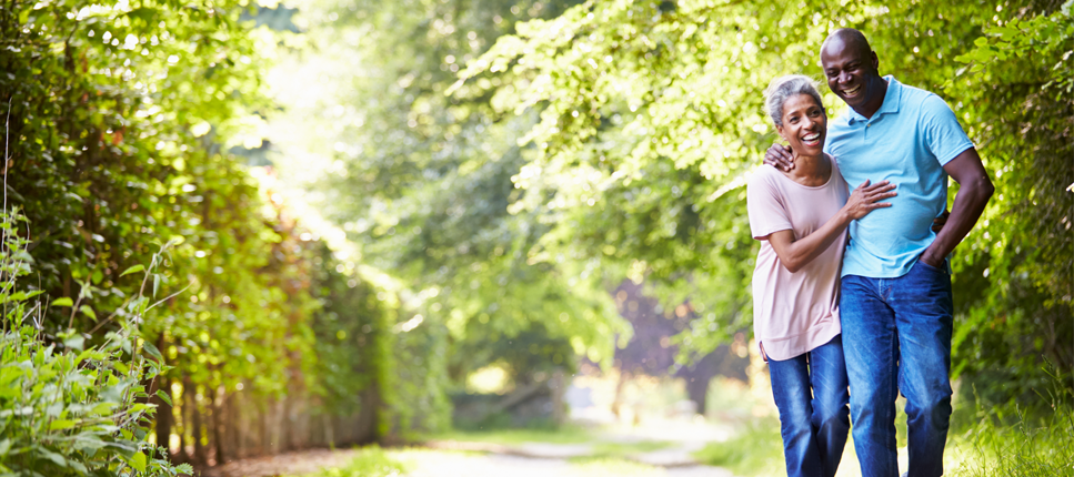 Senior couple walking together in sunlit park path lined with trees during spring season.