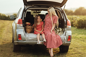 A woman and a young girl in matching red dresses smile at each other while sitting in the open trunk of a silver SUV parked in a golden field