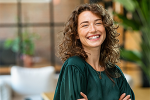 A smiling woman with long curly brown hair wearing a dark green blouse poses with her arms crossed in a brightly lit, modern office setting