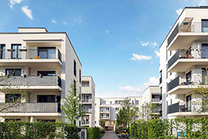 A cluster of modern, white multi-story apartment buildings with balconies surrounds a central courtyard area under a clear blue sky
