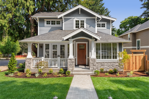 A two-story grey craftsman-style house with white trim and stone accents, featuring a welcoming front porch and a stone walkway through a manicured green lawn