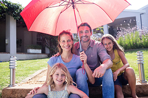 A smiling family of four sits together outdoors on stone steps, sheltered under a large red umbrella on a sunny day