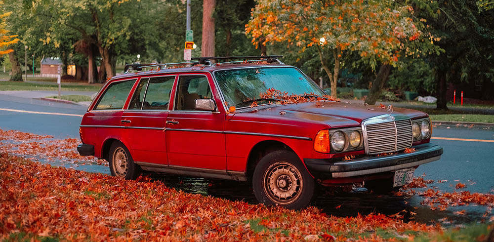 A classic red Mercedes-Benz station wagon is parked along a leaf-strewn roadside during autumn, surrounded by vibrant orange foliage and trees - www.jacaa.co
