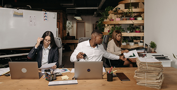 Three office workers sit at a large wooden desk with laptops, documents, and a tall stack of files while collaborating in a modern office space - www.jacaa.co