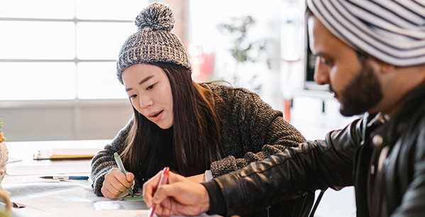A woman wearing a knit pom-pom beanie and a man wearing a striped turban sit next to each other at a desk, focused on writing or drawing together - www.jacaa.co