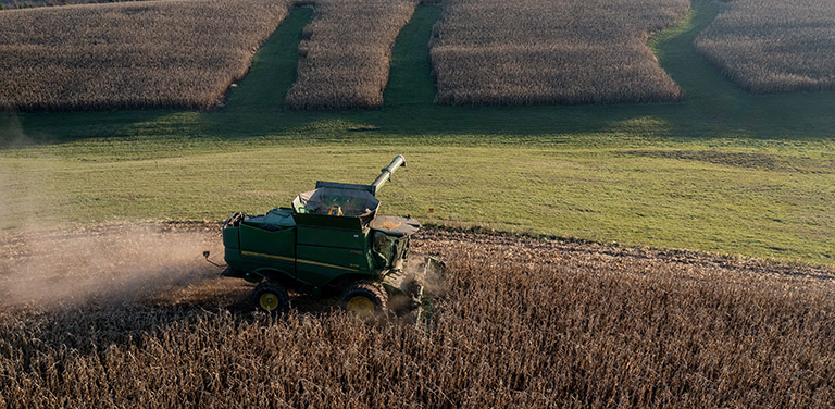 An aerial, high-angle shot captures a green John Deere combine harvester moving across a hillside, kicking up dust as it harvests a dense field of brown, dry crops - www.jacaa.co