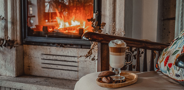 A clear glass mug filled with a layered latte sits on a wooden tray alongside small cookies on an armchair, positioned in front of a warm, roaring fireplace - www.jacaa.co