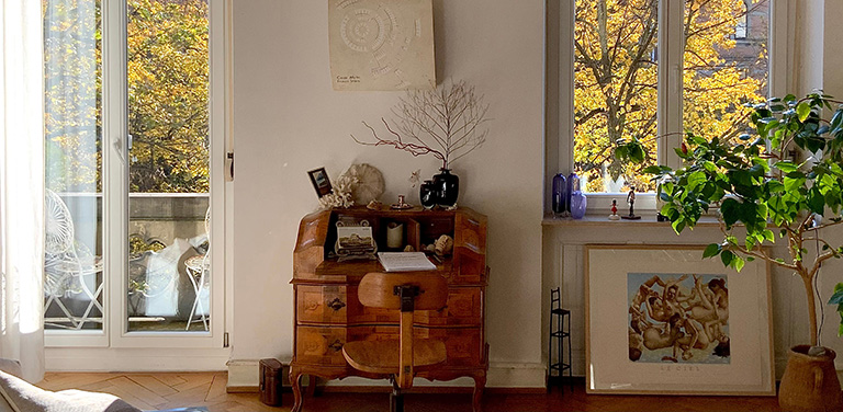 A classic wooden rolltop desk paired with a matching chair sits centered between a glass balcony door and a large window, both showcasing vibrant yellow autumn foliage outside - www.jacaa.co