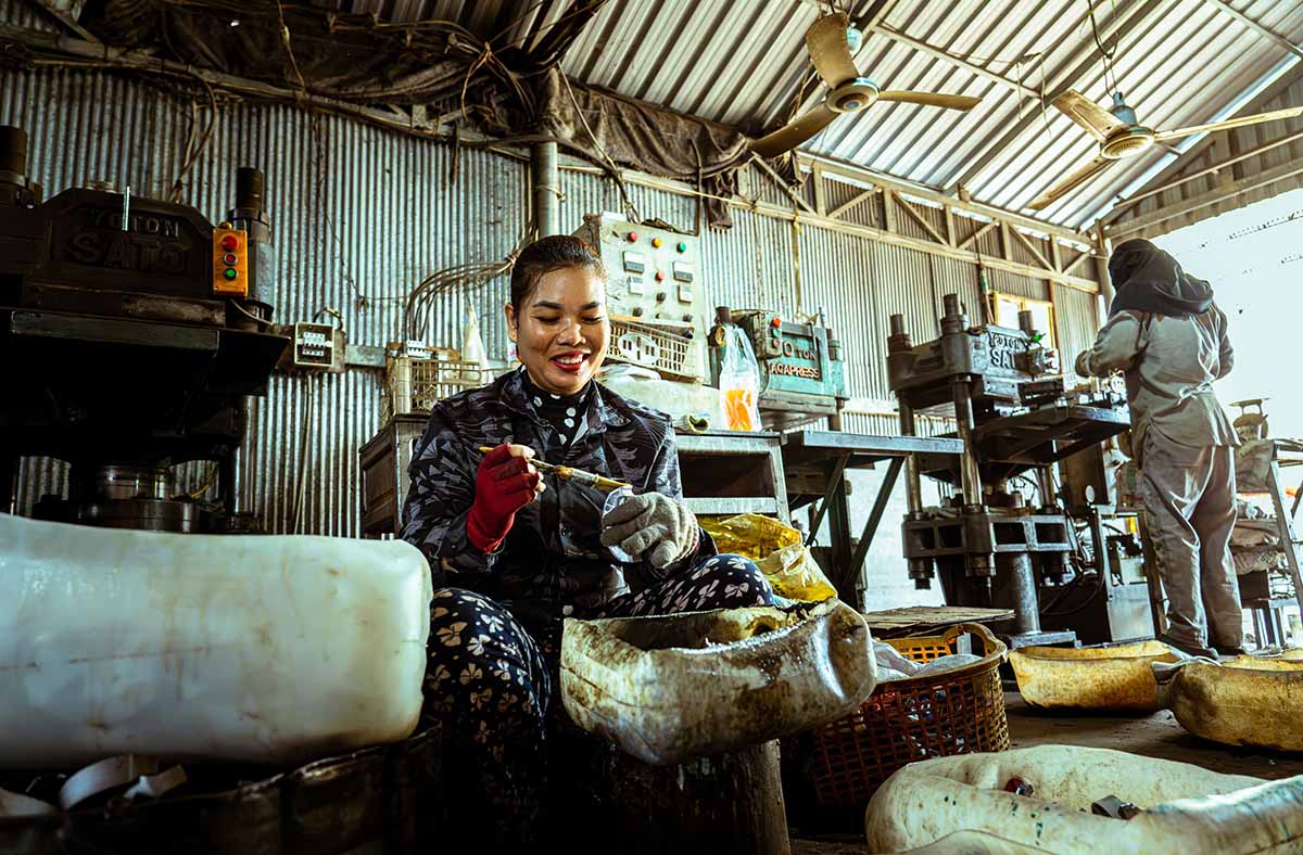 A woman wearing gloves smiles while sitting and working with a tool inside an industrial workshop with machinery and another worker in the background - www.jacaa.co